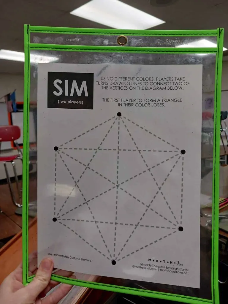hand holding up template for game of sim in dry erase pocket in high school math classroom 